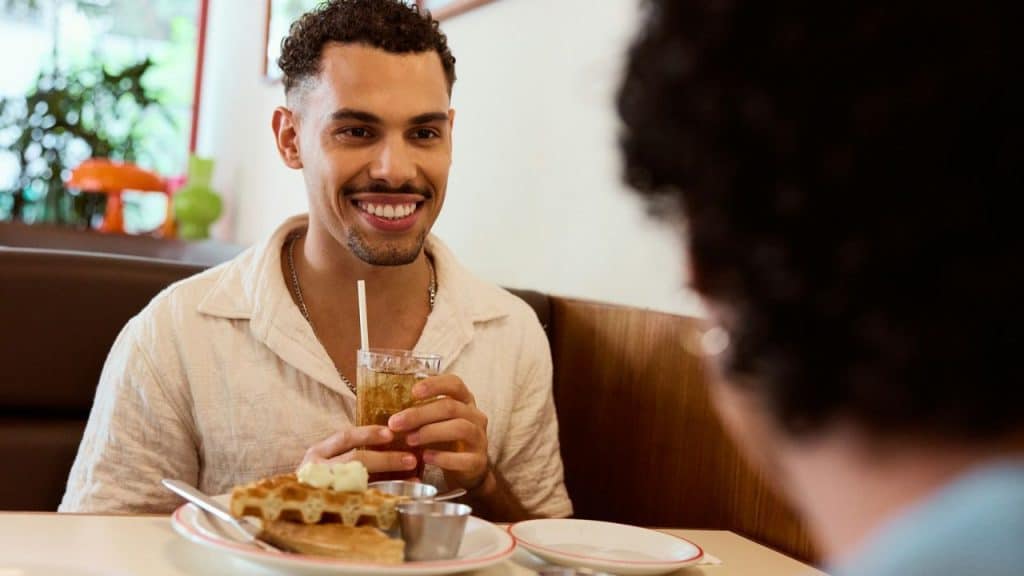 Smiling man holding a drink and looking at another person across a diner table.