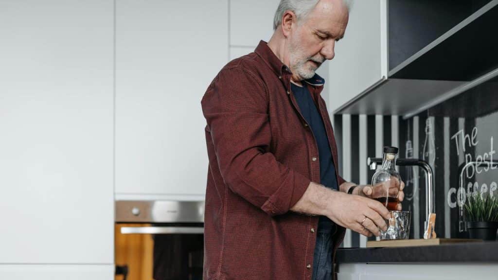 An older man pouring a drink into a glass in a kitchen.
