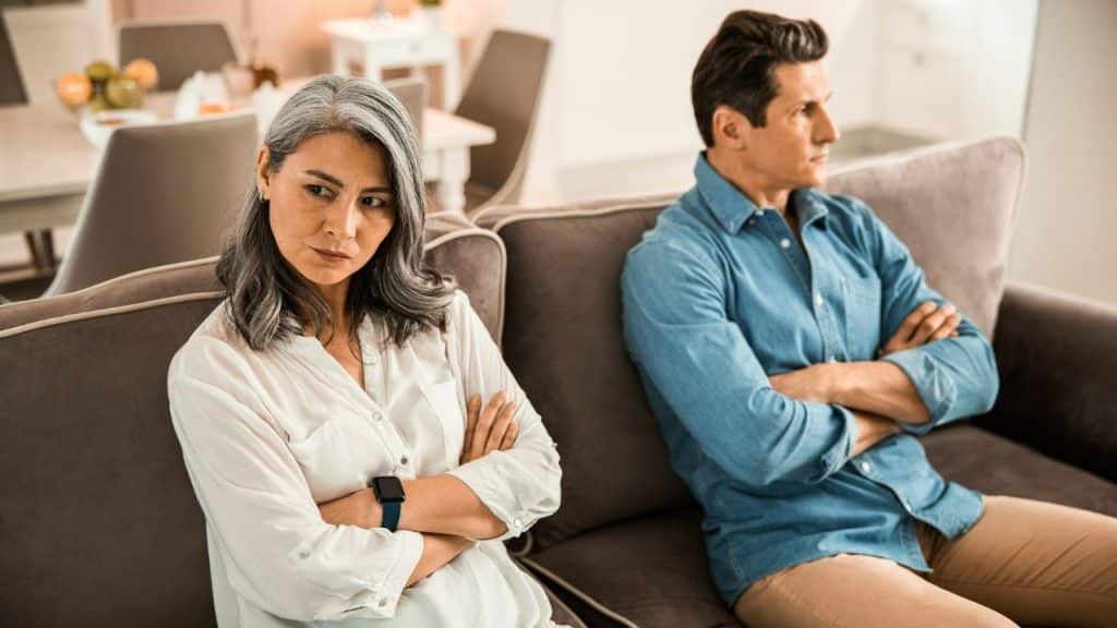 A serious couple sits on a couch with their arms crossed, facing away from each other.