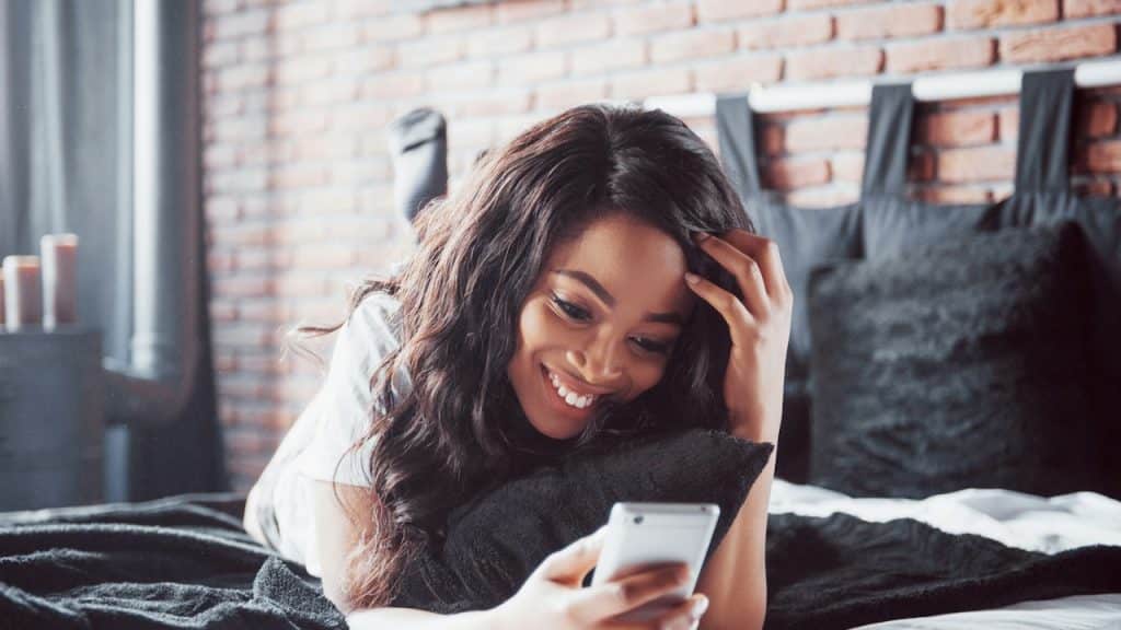 A smiling young woman with long, dark hair is lying on a bed and looking at her smartphone.