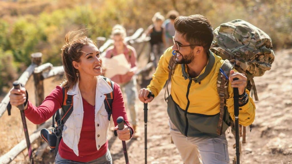 A smiling couple with backpacks and trekking poles is hiking up a sunny dirt trail with other people behind them.