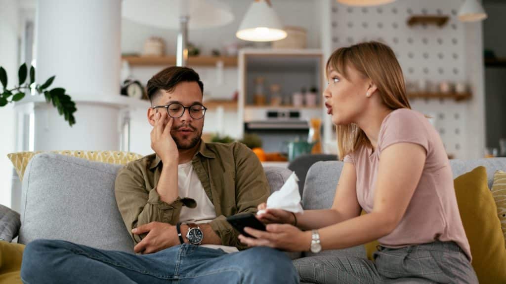 A couple sitting on a couch, with the woman arguing and the man looking tired.