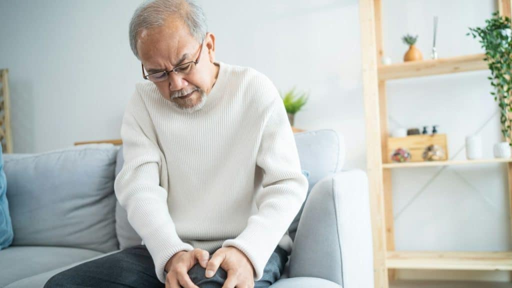 Elderly man in a white sweater sitting on a couch grasping his knee in pain.