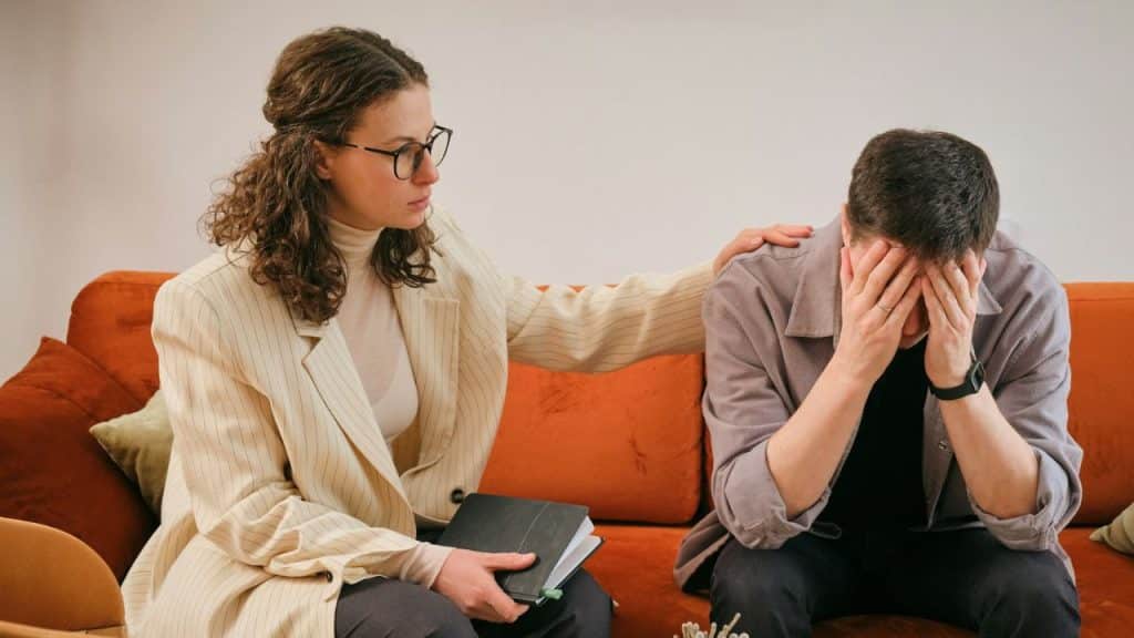 Woman comforting a crying man by placing her hand on his shoulder in a therapy session.