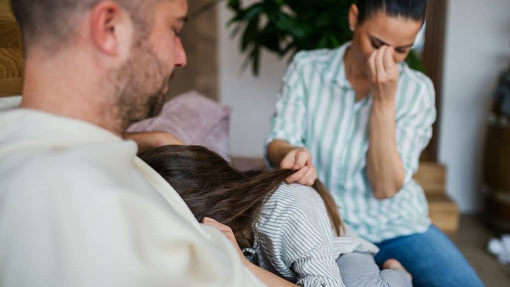 A distraught family scene where a crying child is comforted by a man on a couch, and a woman sits behind them with her hand on her face.