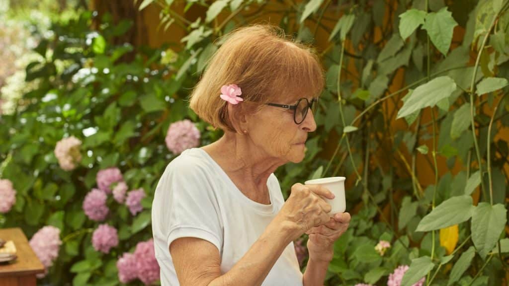 A woman wearing glasses sipping coffee from a cup in a greenhouse.