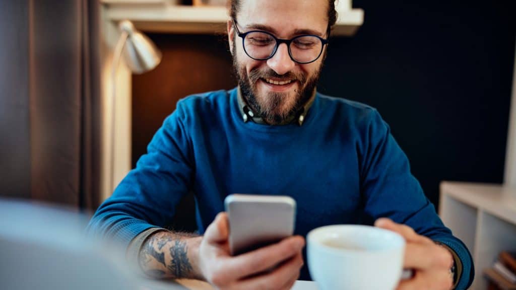 A bearded man with glasses and a blue sweater smiles while looking at his smartphone, holding a white coffee mug in his other hand at a desk.