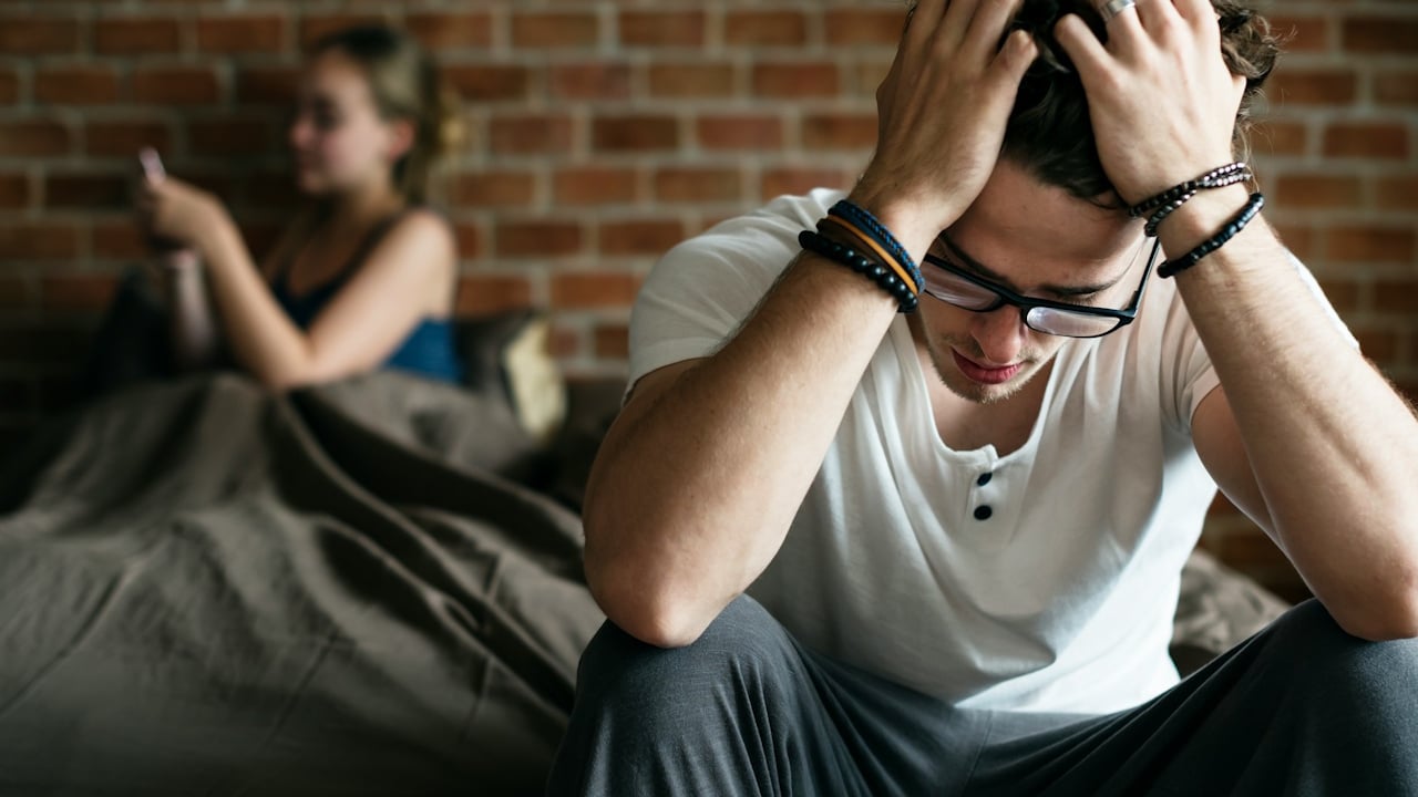 A woman is texting her ex while her worried husband is sitting holding his head with his hands.