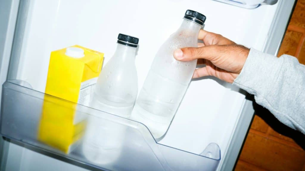 A person's arm reaching into a refrigerator to grab a frosty glass bottle of water.