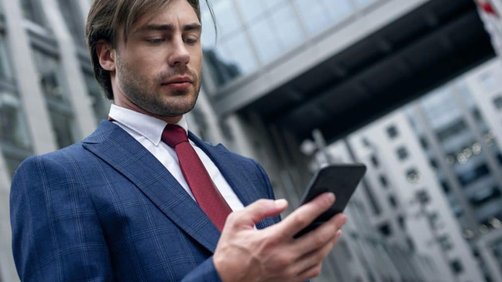 Man in a blue plaid suit and a red tie looking at his smartphone outside a modern building.