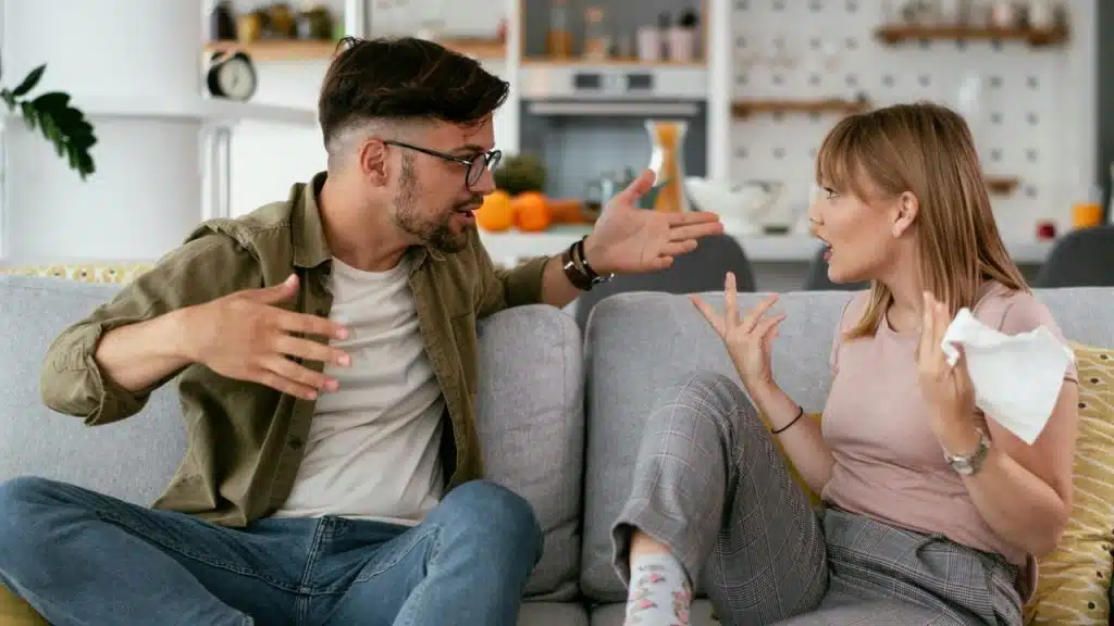 A man and woman on a couch are arguing and gesturing dramatically at each other.