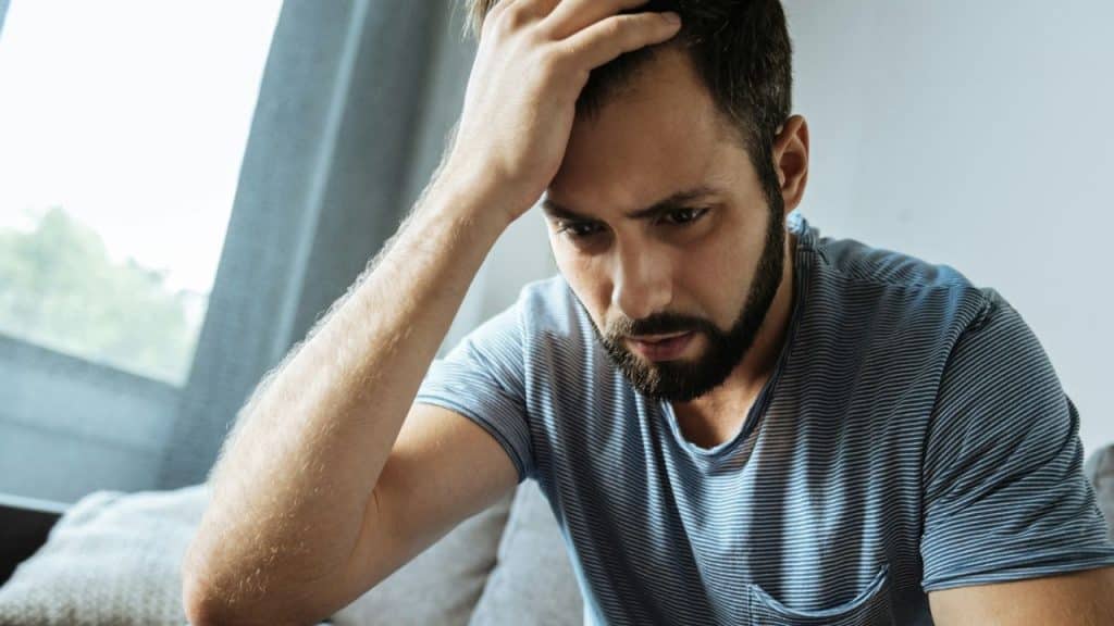 A distressed, bearded man in a striped shirt holds his head with one hand.