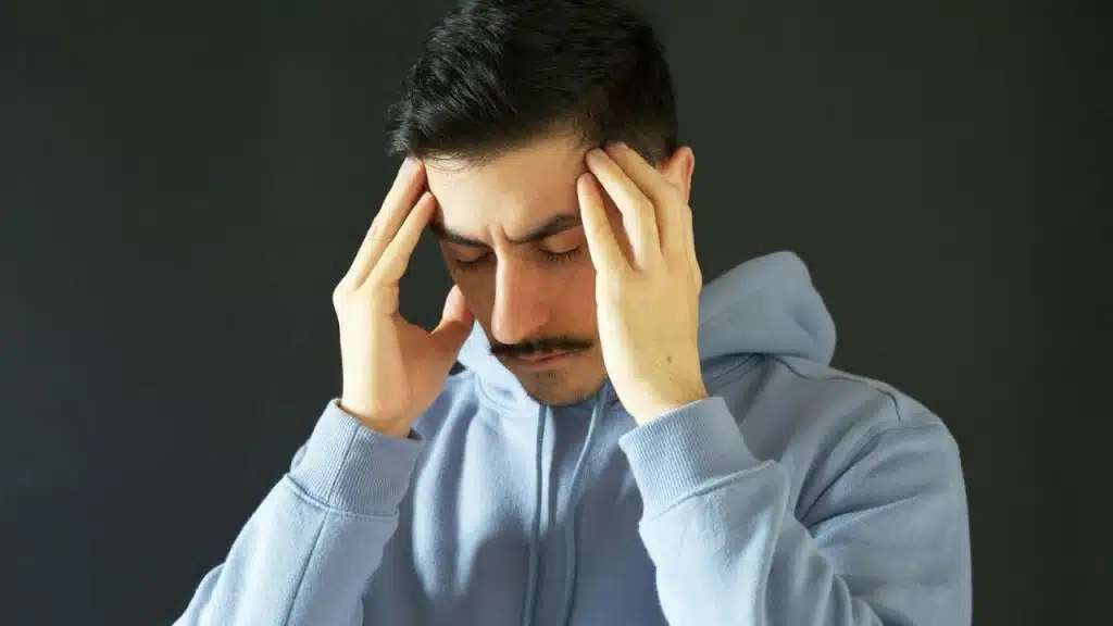 A man with a mustache, wearing a blue hoodie, holds his temples with his eyes closed.