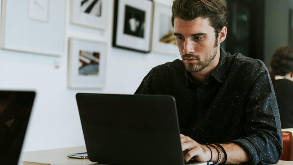 Concentrated man working on a black laptop at a desk with framed art on the wall.