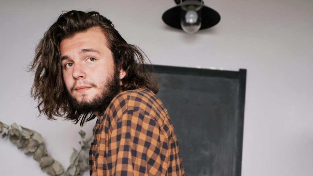 Young man with long, wavy hair and a beard wearing a plaid shirt looking at the camera.