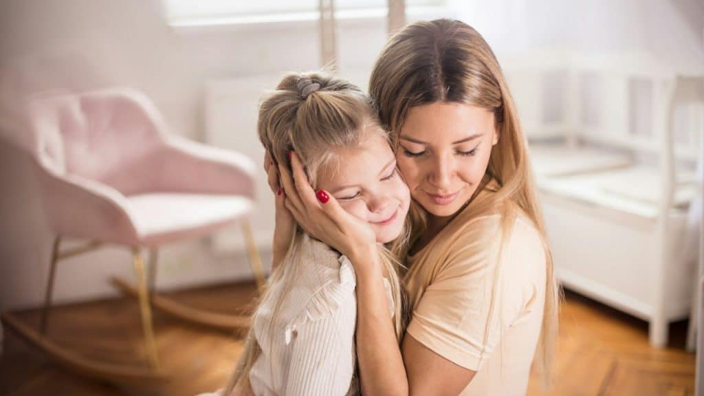 Woman in a beige shirt embracing a smiling young girl with long blonde hair.
