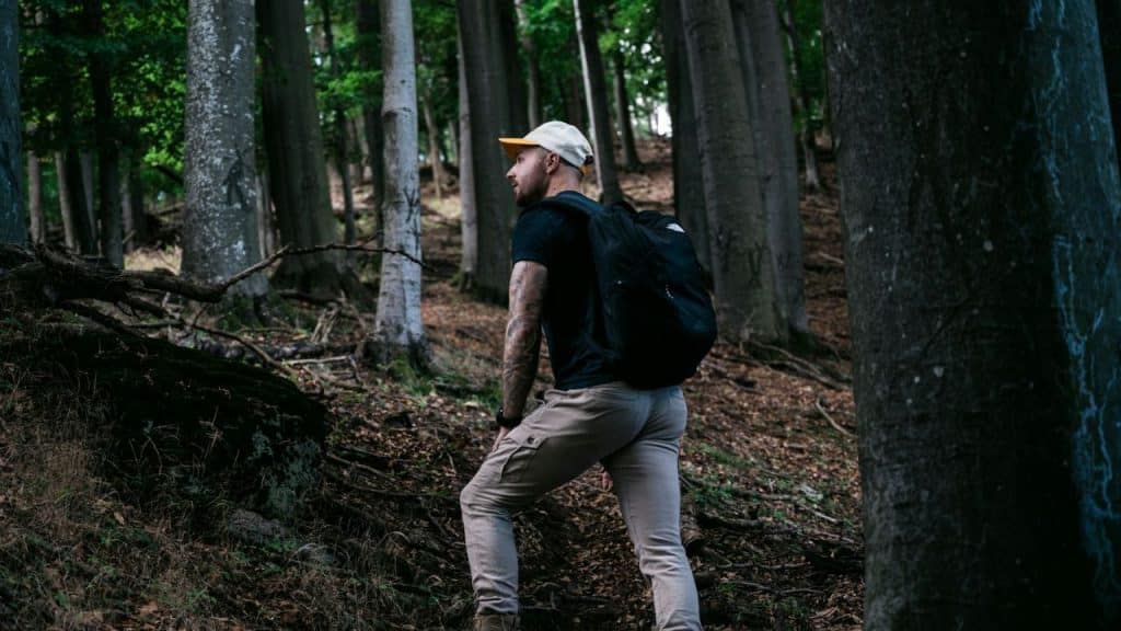Man wearing a hat and backpack hiking up a dirt path in a dense forest.