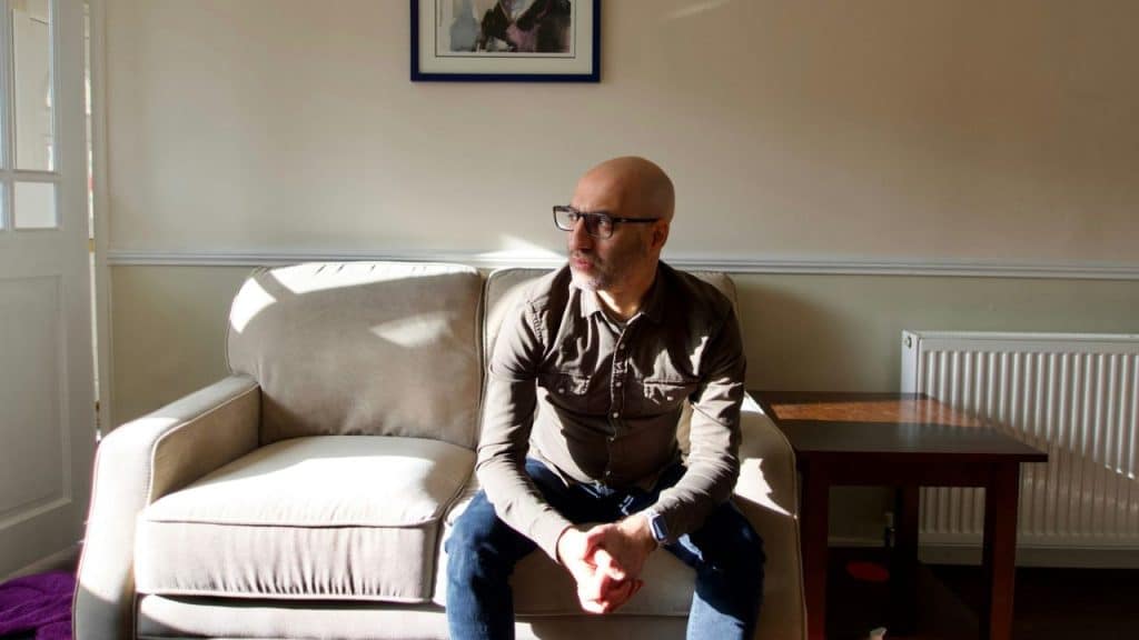 Bald man with glasses sitting on a light-colored couch in a room with strong sunlight.