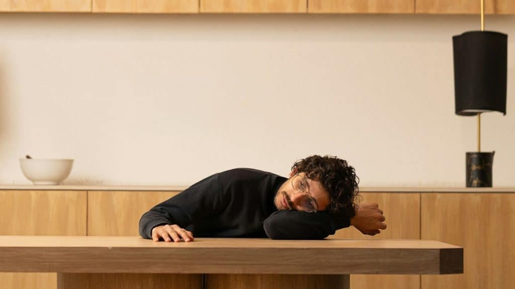 Man resting his head on a wooden table in a brightly lit modern kitchen.