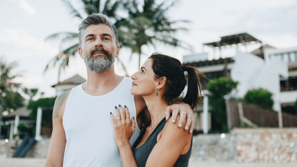 Man with beard and woman embracing outdoors against tropical resort background.