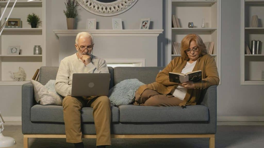 An elderly couple relaxing on a couch reading a book and working on a laptop.