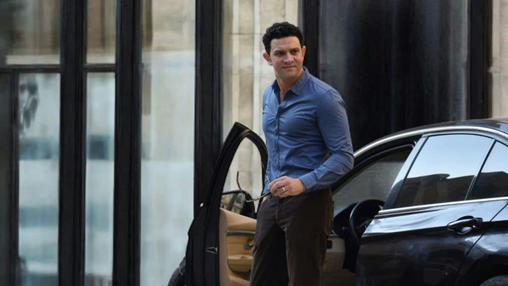 Man in a blue shirt and brown pants getting out of a black car.
