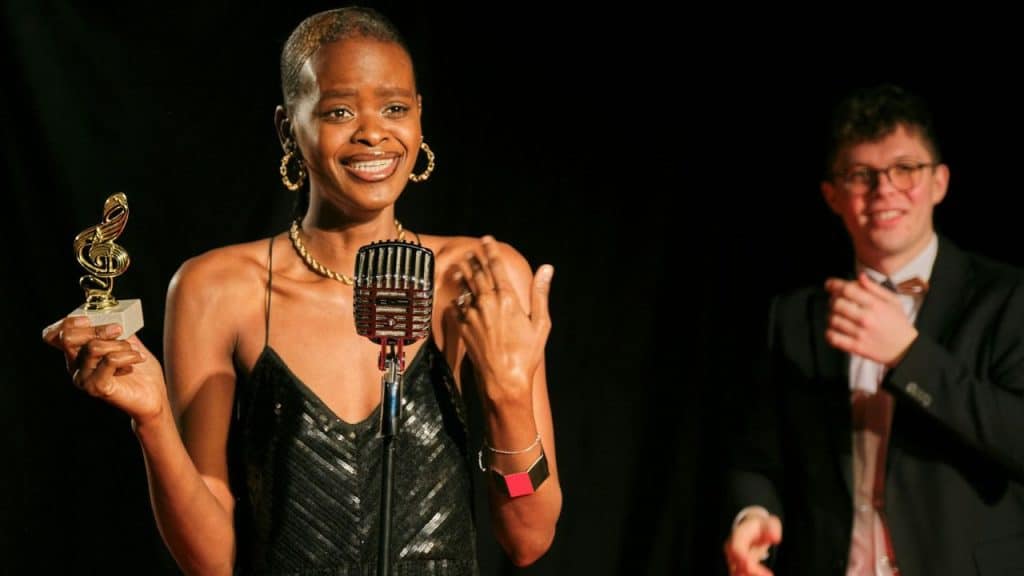 A smiling woman holding a treble clef award stands at a vintage microphone on a stage.