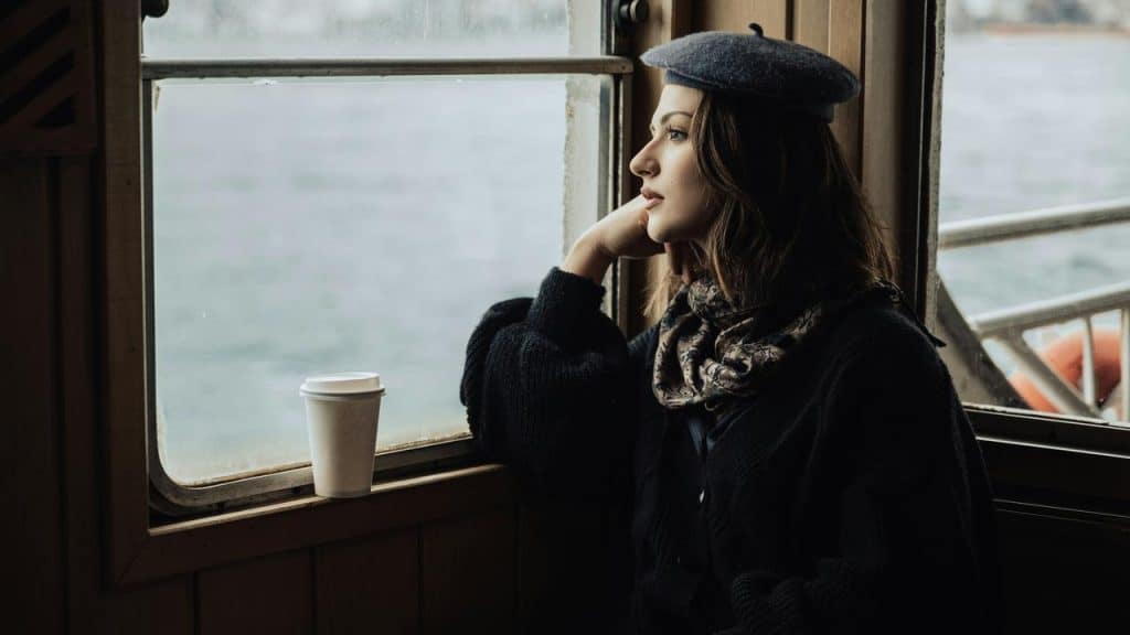A woman sitting by a window with a cup of coffee, looking outside.