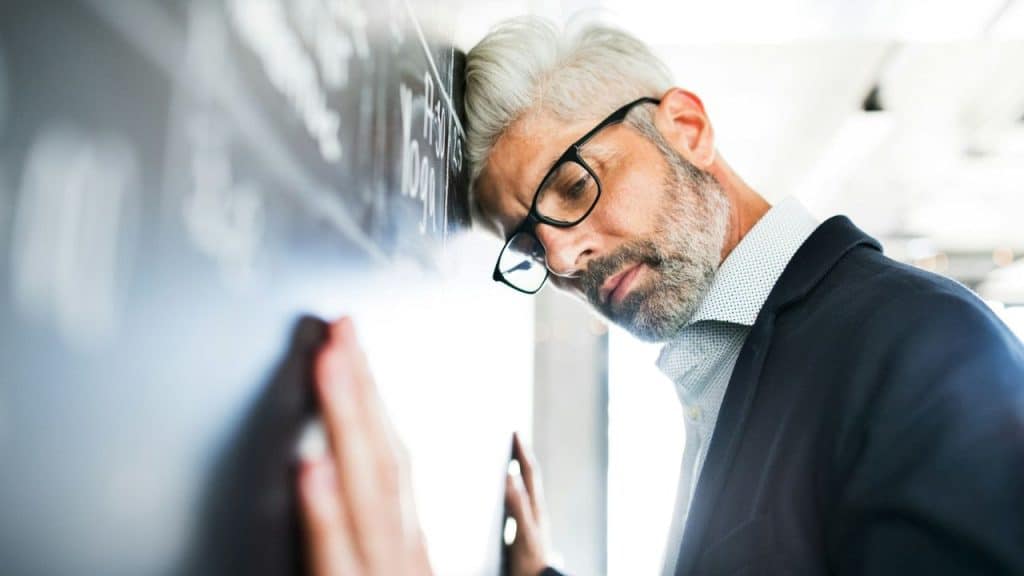 Middle-aged man in glasses leaning his head against a dark chalk wall with writing.