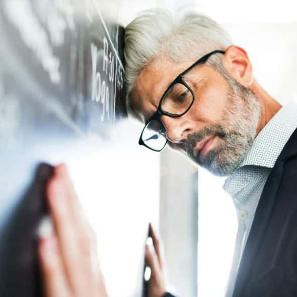 Middle-aged man in glasses leaning his head against a dark chalk wall with writing.