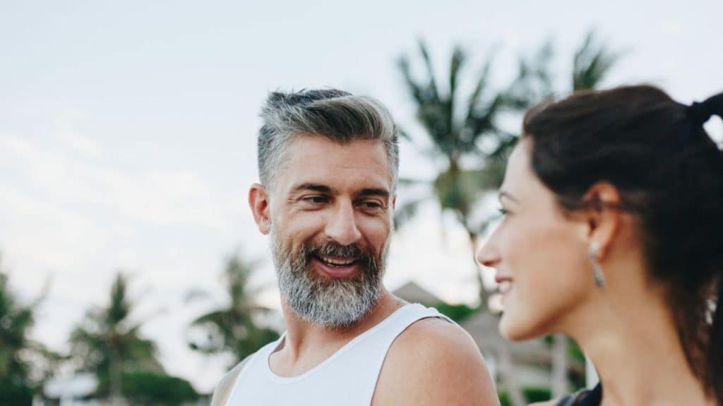 Middle-aged man with a gray beard smiling at a woman outdoors with palm trees.