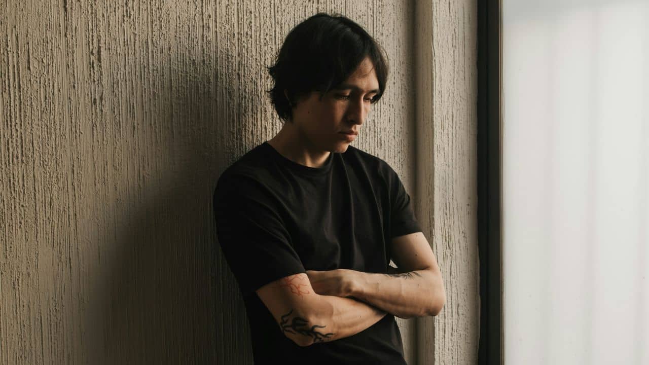 Young man with black hair and tattoos standing, looking down with crossed arms.