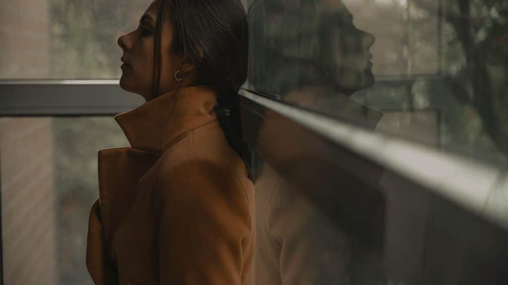 A person in a brown coat leans against a glass wall, reflected in the window.