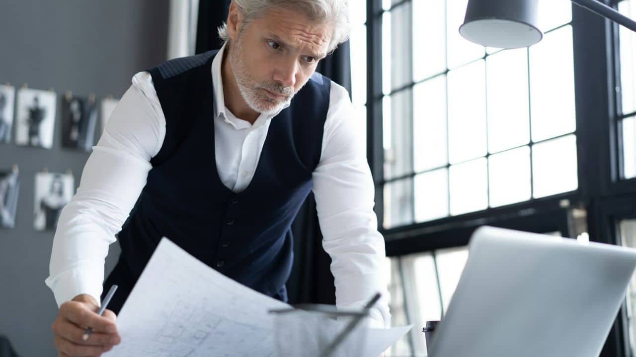 Middle-aged man in a vest and white shirt intently studying papers near a laptop.
