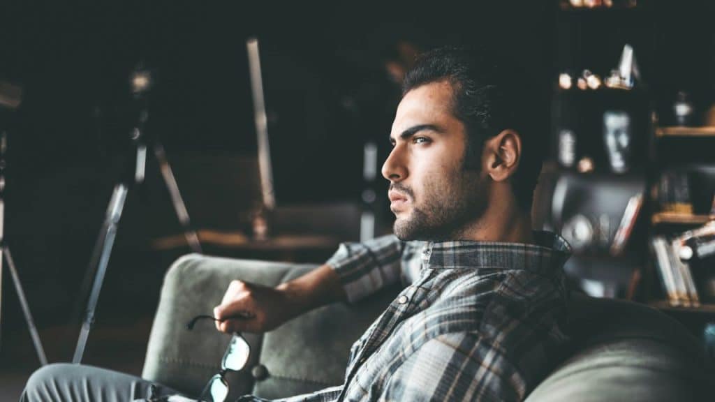 Young man in a plaid shirt sitting in a chair, holding glasses and looking away.