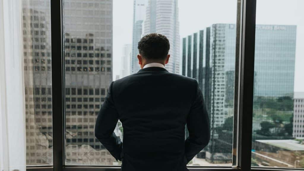 Man in a suit seen from the back, looking out a large window at skyscrapers.