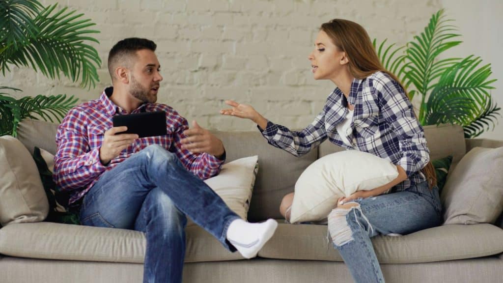 A woman talks and gestures to a man holding a small tablet on a couch.