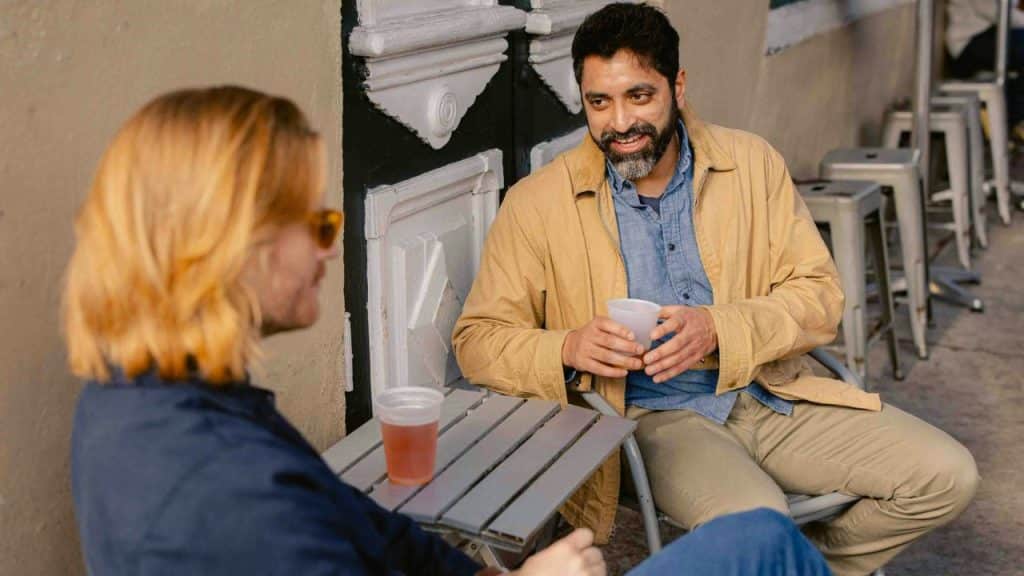 Two people sitting at a small outdoor table, talking and holding plastic cups.
