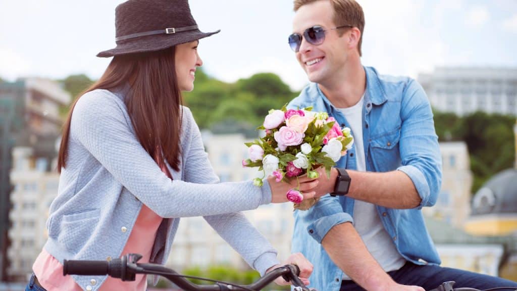 Smiling couple on bicycles, the man presenting a bouquet of flowers to the woman.