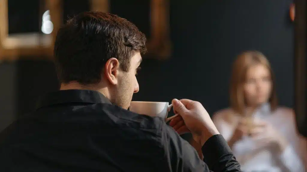 A man in a black shirt drinking coffee with a woman sitting in the background.