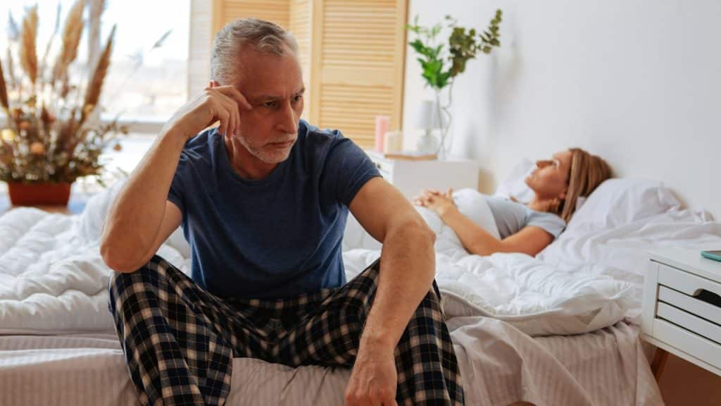 An Upset man sitting on bed with his head in his hand, while a woman lying turned away from him in the background.