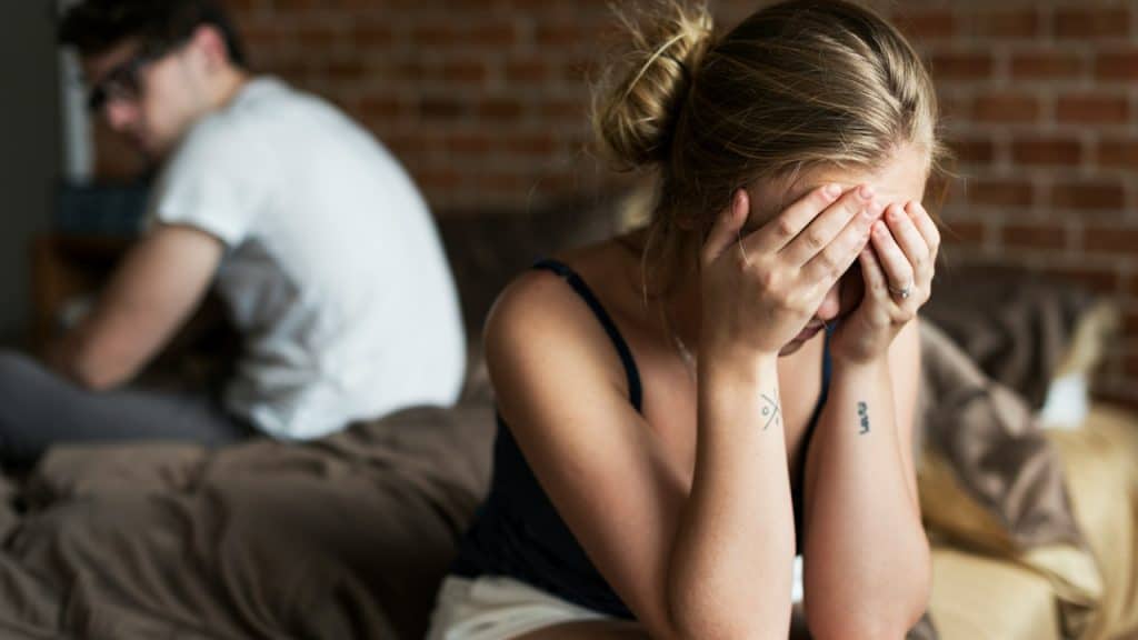 A couple sitting on opposite sides of a bed while a woman cries with her face hidden in her hands and the man looks at her from the side of his vision.