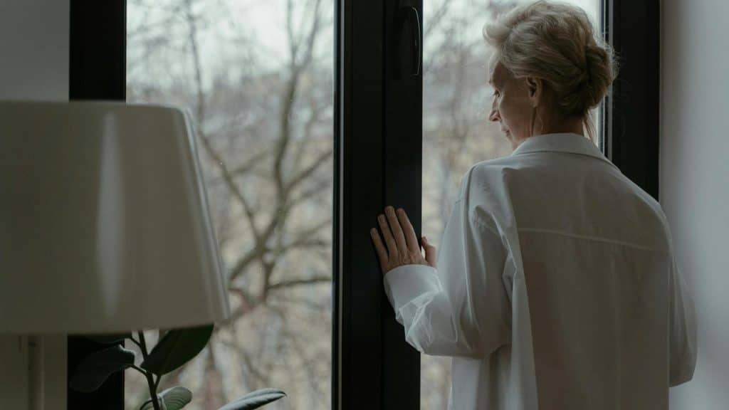An older woman in a white shirt looks out a window.