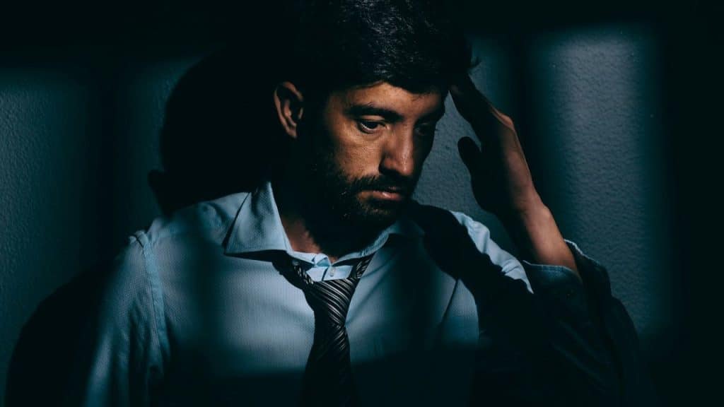 A man in a shirt and tie with a beard is holding his head, looking down in a dark room.