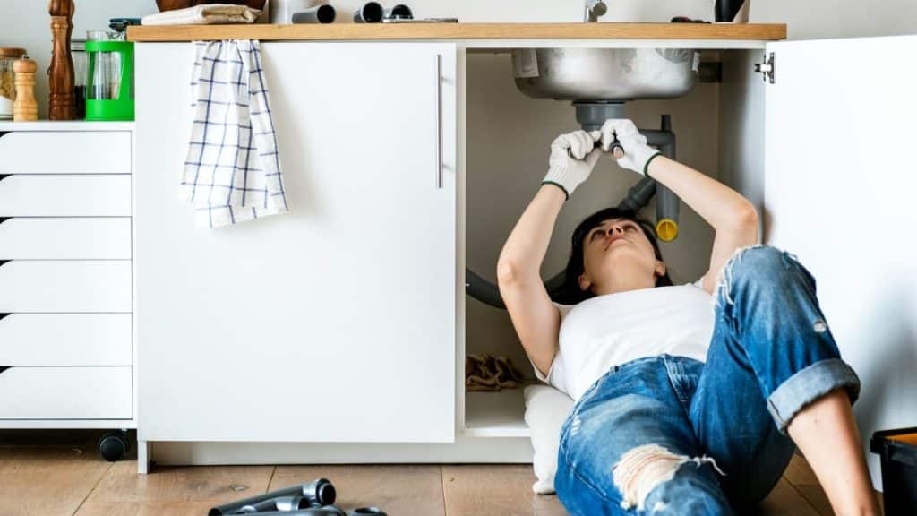 A woman in a white shirt and ripped jeans lies under a kitchen sink, fixing a pipe.