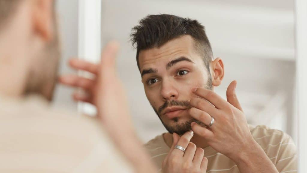 A man looks intensely into a mirror, touching his face with his fingers.