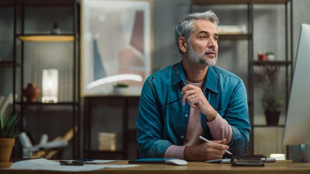 Middle-aged man in a denim shirt sits at a desk, looking thoughtful with a stylus.