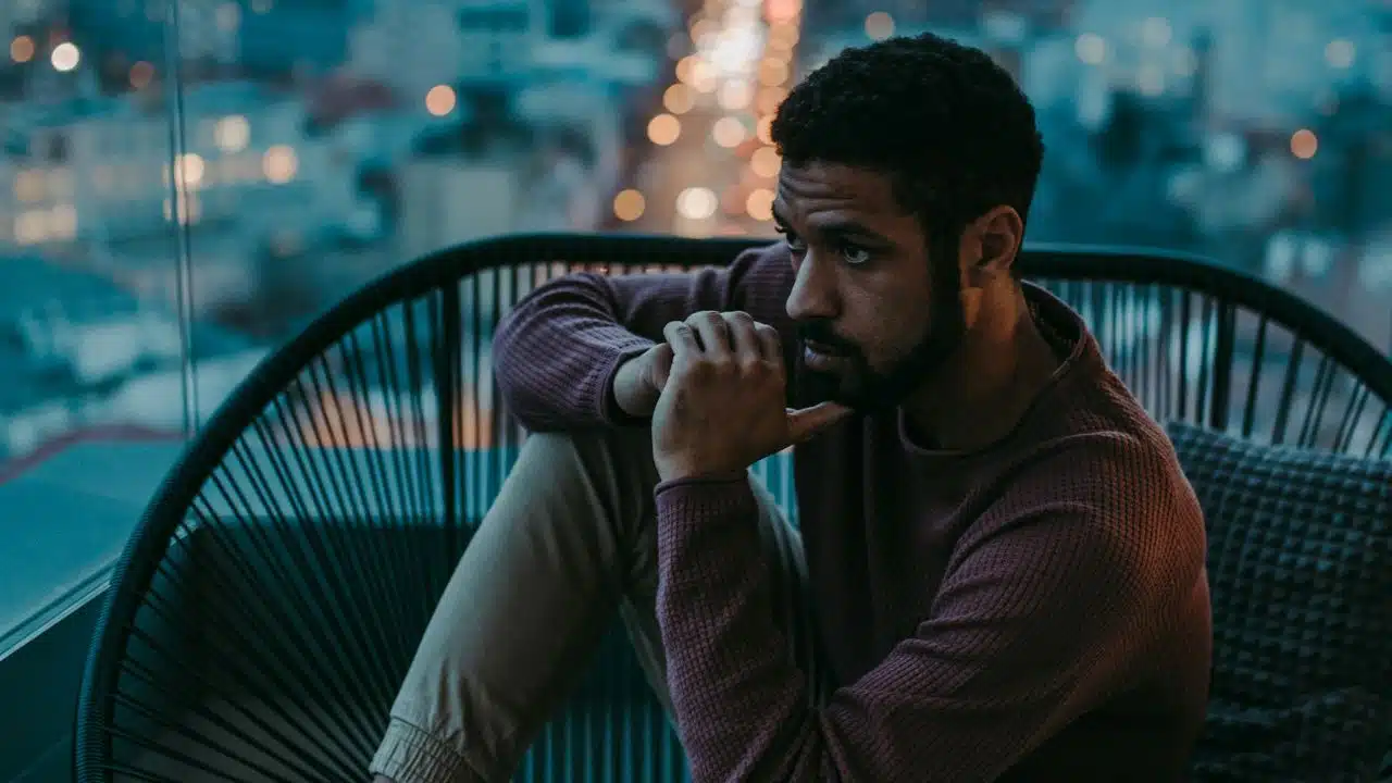 Man sitting alone, looking out at blurred city lights at night, appearing thoughtful.