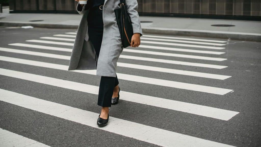 A person in a gray coat walking across a crosswalk with coffee and a bag.