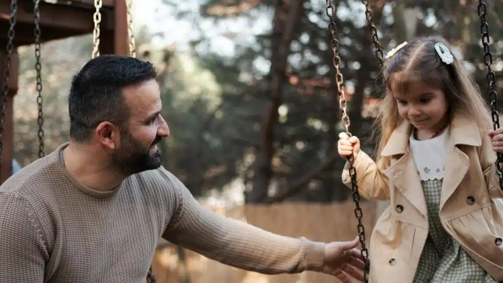 A man smiles at a little girl on a swing in an outdoor playground.